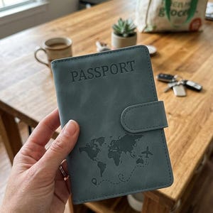 May include: A slate blue passport holder featuring the word PASSPORT embossed at the top. The cover displays a dark gray world map graphic with a dotted flight path and a small airplane icon. It includes a side strap with a magnetic closure.