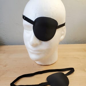 May include: A solid black eye patch featuring a smooth, rounded, opaque shield and an adjustable black elastic strap with a metal slider. One eye patch is displayed on a white mannequin head, while a second identical eye patch lies flat below.