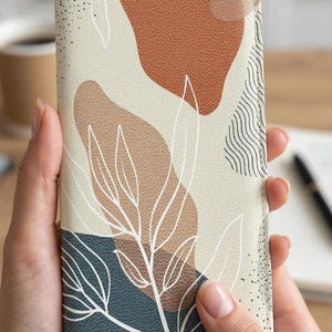 May include: A wallet with an abstract design featuring earth-tone shapes and white leaf outlines. The wallet has a textured surface and a color palette of beige, brown, and teal. The design is modern and artistic.