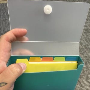 May include: A teal file box with a silver lid held open, revealing yellow file folders with colorful tabs. The box is held by a hand, and a white circular magnet secures the lid.
