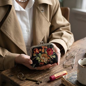 May include: A small black coin purse with a silver metal kiss-lock clasp, featuring an embroidered floral pattern in shades of red, orange, and green. The purse has a structured rectangular shape and is held in place, showcasing the detailed needlework.