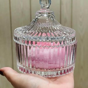 May include: A clear, decorative glass jar with a lid, featuring a ribbed design. The jar contains a pink item, possibly cotton balls or a similar product. The jar is held in a hand.