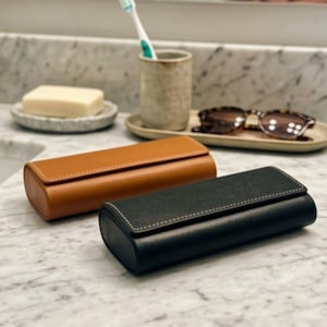 May include: Two rectangular eyeglass cases, one in tan and one in black, featuring a smooth finish and visible white stitching along the edges. The cases have a flap closure and a rounded profile, positioned on a white marble surface.