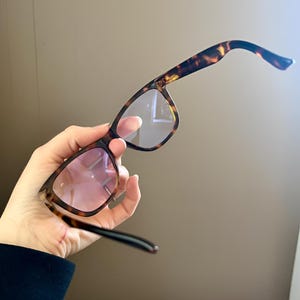 May include: Tortoiseshell-patterned eyeglasses featuring a classic frame design with dark brown and amber mottled coloring. The lenses have a subtle pinkish tint, and the temples are curved at the ends for a secure fit around the ears.