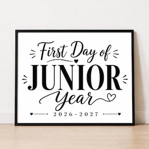 May include: A rectangular black frame containing a white sign with black text that reads First Day of JUNIOR Year 2026-2027. The design features decorative flourishes, small hearts, and lines surrounding the bold, centered lettering.