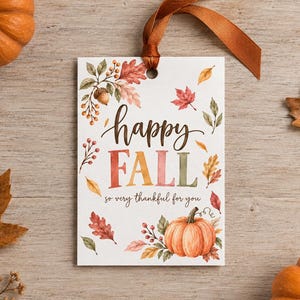 May include: A rectangular white gift tag with a brown ribbon, featuring watercolour illustrations of autumn leaves, acorns, and a pumpkin. The text reads "happy FALL" in brown and multicoloured letters, followed by the script text "so very thankful for you."