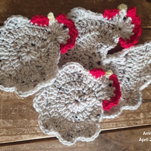 Puede incluir: Posavasos hechos a mano en crochet con forma de gallina. Los posavasos son grises con motas de color, con detalles de cresta y barbilla rojos. Los posavasos están dispuestos sobre una superficie de madera.