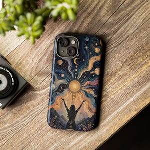 Könnte beinhalten: Eine Handyhülle mit einem kosmischen Design, das eine Sonne, Planeten und eine Silhouette einer Person mit erhobenen Armen zeigt. Die Hülle hat einen dunkelblauen Hintergrund mit goldenen und orangenen Akzenten. Die Handyhülle liegt auf einer Holzoberfläche.