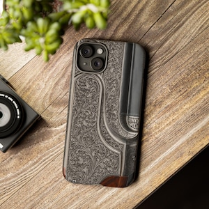 May include: A phone case featuring an intricate floral engraving in grey and silver tones. The case incorporates a dark grey, leather-effect section and a small, dark wood detail at the base. It rests on a wooden surface.