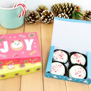 May include: Two gift boxes with festive designs. The larger box is pink with a hedgehog and the word "JOY" printed on it. The smaller box is yellow with a hedgehog design. The smaller box is open and contains four white chocolate peppermint cookies.