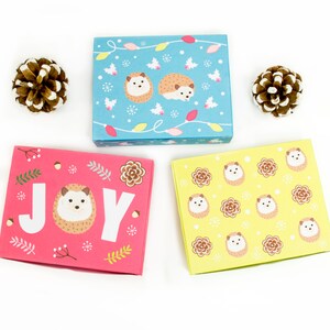 May include: Three gift boxes with a hedgehog theme. The first box is blue with a white snowflake pattern and hedgehogs with Christmas lights. The second box is pink with a white snowflake pattern and the word "JOY" in white letters. The third box is yellow with a white snowflake pattern and hedgehogs.