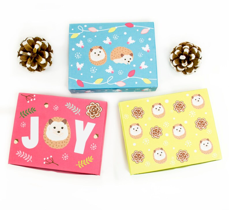 May include: Three gift boxes with a hedgehog theme. The first box is blue with a white snowflake pattern and hedgehogs with Christmas lights. The second box is pink with a white snowflake pattern and the word "JOY" in white letters. The third box is yellow with a white snowflake pattern and hedgehogs.