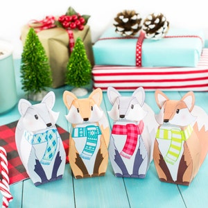 May include: Four papercraft fox ornaments with different colored scarves. The foxes are standing on a blue wooden surface. The foxes are wearing scarves with a snowflake, plaid, green and white stripes, and a red and white plaid pattern.