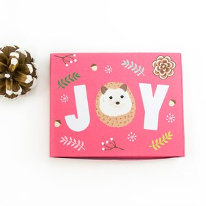 May include: A pink gift box with a hedgehog and the word "JOY" printed on it. The box is decorated with small green leaves, white snowflakes, and brown acorns.