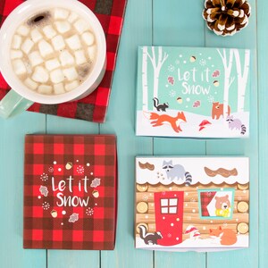以下が含まれることがあります： 「Let it Snow」と書かれた赤と黒の格子縞のギフトボックスが2つ。1つのボックスには、キツネ、アライグマ、クマが描かれた冬のシーンが描かれている。もう1つのボックスには、屋根にアライグマ、窓にクマがいる小屋が描かれている。