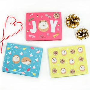 May include: Three gift boxes with a Christmas theme. The boxes are decorated with hedgehogs and the word "JOY". One box is pink with white snowflakes, one is blue with white lights, and one is yellow with brown hedgehogs.