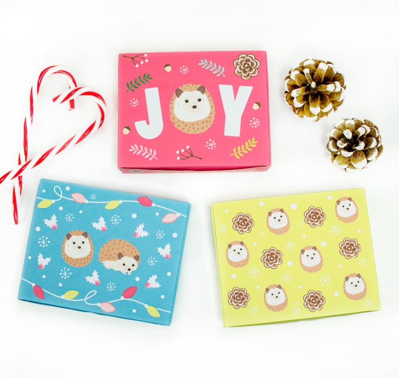 May include: Three gift boxes with a Christmas theme. The boxes are decorated with hedgehogs and the word "JOY". One box is pink with white snowflakes, one is blue with white lights, and one is yellow with brown hedgehogs.