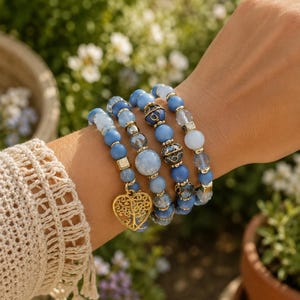 May include: A set of four stacked beaded bracelets featuring various shades of blue, translucent, and opaque beads. The bracelets include gold-toned spacers and a prominent gold-toned heart-shaped charm with an intricate tree of life cutout design.