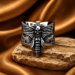 May include: A silver-toned metal ring featuring a detailed moth design with a skull motif centred on the thorax. The wings are spread wide with intricate line textures, and the body of the moth extends vertically along the band of the ring.