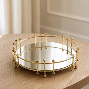 Könnte beinhalten: Ein rundes Tablett mit einer reflektierenden Spiegeloberfläche und einem goldfarbenen Metallrahmen. Der Rahmen besteht aus einer dünnen kreisförmigen Schiene, die von vertikalen Metallstäben mit kleinen Kugeln an den Enden gestützt wird.