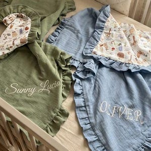 May include: Two muslin baby blankets with ruffled edges, one sage green embroidered with Sunny Lucia and one dusty blue embroidered with OLIVER. Both feature a patterned underside with illustrations of woodland animals and plants.
