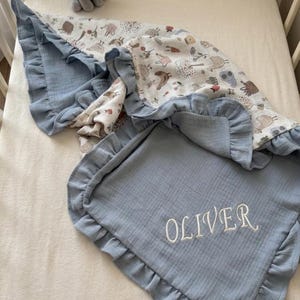 May include: A blue muslin baby blanket with a ruffled edge and the name OLIVER embroidered in white thread. The blanket features a patterned side with illustrations of animals and trees, and a small gray plush elephant toy sits nearby.
