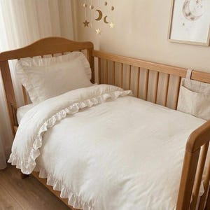Könnte beinhalten: Ein hölzernes Kinderbett mit einem weißen, gerüschten Bettbezug und einem passenden gerüschten Kissen. Über dem Bett hängt ein Mobile mit goldfarbenen Mond- und Sternformen, und an der Seitenreling ist eine Aufbewahrungstasche aus beigem Stoff.