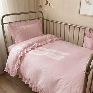 Könnte beinhalten: Ein Metallbett mit bronzefarbenem Finish und hellrosa Bettwäsche. Das Set besteht aus einem Kissen mit Rüschenrand und einem passenden Bettbezug mit Rüschenbesatz. Eine kleine rosa Stofftasche hängt an der Seite des Bettgitters.