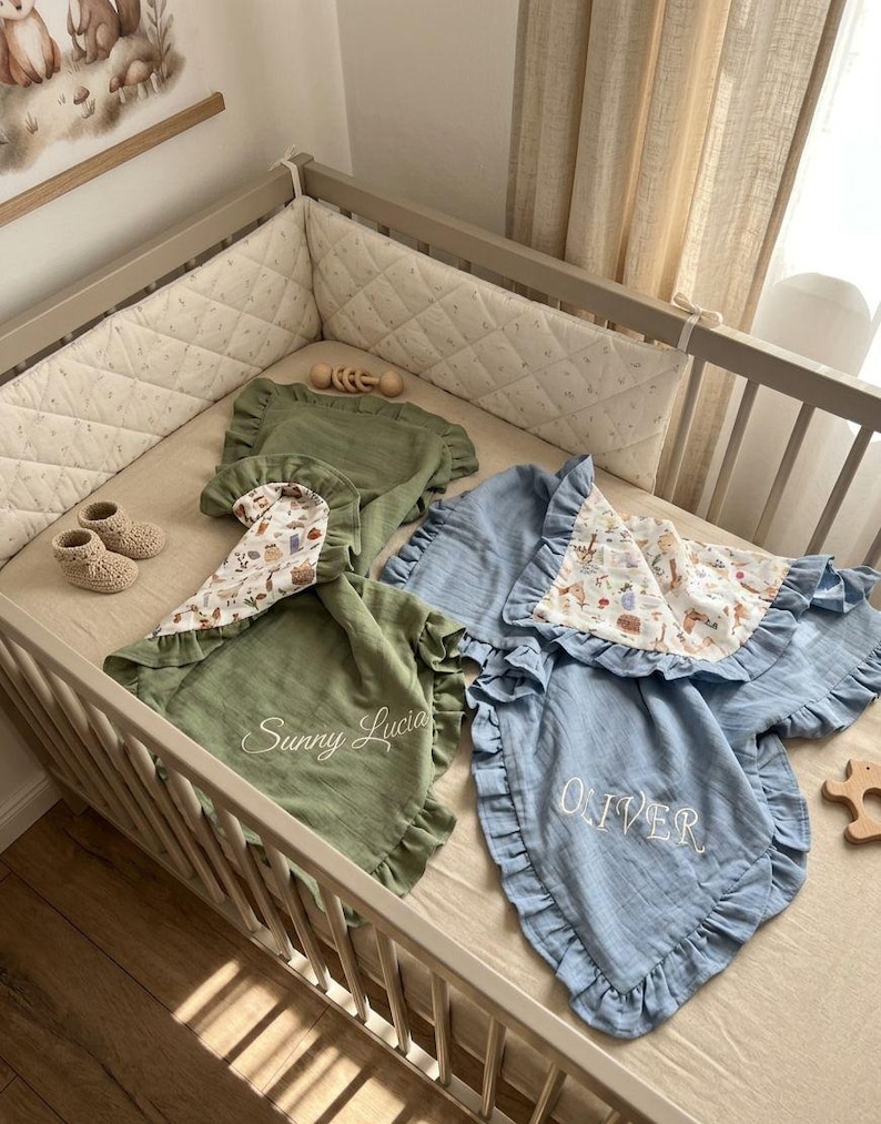 May include: Two muslin baby blankets with ruffled edges, one sage green embroidered with Sunny Lucia and one dusty blue embroidered with OLIVER. Both feature a patterned underside with illustrations of woodland animals and plants.