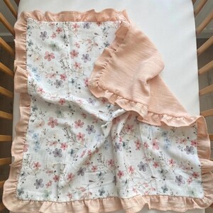 May include: A rectangular baby blanket featuring a white background with a delicate floral pattern in shades of pink, blue, and grey. The blanket is bordered by a solid peach-colored ruffled trim that extends along all four edges of the fabric.
