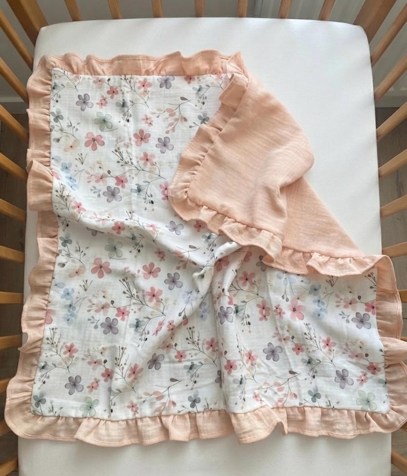 May include: A rectangular baby blanket featuring a white background with a delicate floral pattern in shades of pink, blue, and grey. The blanket is bordered by a solid peach-colored ruffled trim that extends along all four edges of the fabric.