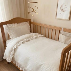 Può includere: Una culla in legno con un copripiumino bianco con volant e un cuscino abbinato con volant. Una giostrina sospesa con forme di luna e stella color oro è appesa sopra il letto e una tasca portaoggetti in tessuto beige pende dalla sponda.