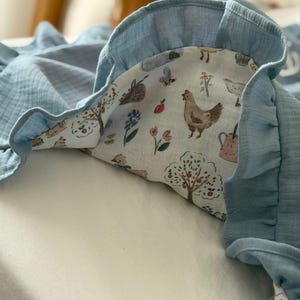 May include: A soft, blue muslin blanket featuring a ruffled edge and a white underside printed with a colorful pattern of chickens, apple trees, flowers, bees, and watering cans. The fabric has a crinkled texture throughout the entire piece.