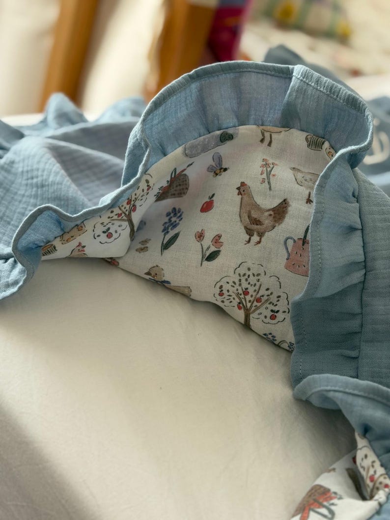 May include: A soft, blue muslin blanket featuring a ruffled edge and a white underside printed with a colorful pattern of chickens, apple trees, flowers, bees, and watering cans. The fabric has a crinkled texture throughout the entire piece.