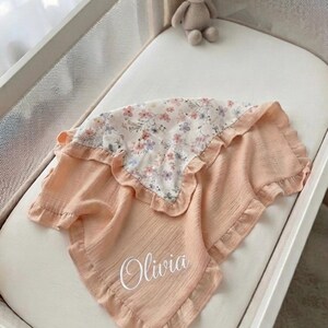 May include: A peach-colored muslin baby blanket with a ruffled edge and a floral patterned section featuring small pink and purple flowers. The blanket is personalized with the name Olivia in white script lettering on the solid peach fabric.
