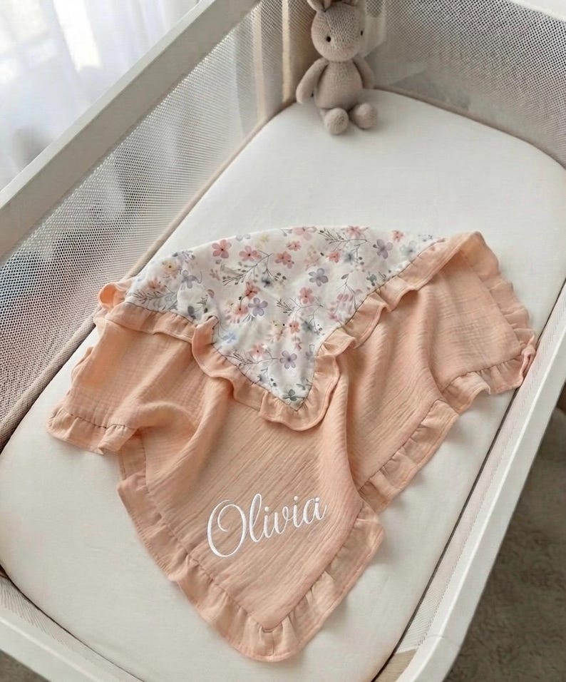 May include: A peach-colored muslin baby blanket with a ruffled edge and a floral patterned section featuring small pink and purple flowers. The blanket is personalized with the name Olivia in white script lettering on the solid peach fabric.