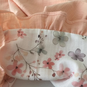 May include: A soft, textured fabric featuring a solid peach-colored ruffled border attached to a white panel. The white section is decorated with a delicate floral pattern consisting of small pink and grey flowers on thin, dark brown stems.