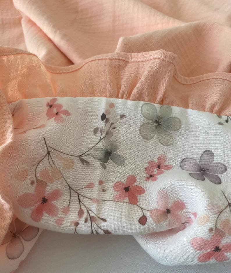 May include: A soft, textured fabric featuring a solid peach-colored ruffled border attached to a white panel. The white section is decorated with a delicate floral pattern consisting of small pink and grey flowers on thin, dark brown stems.
