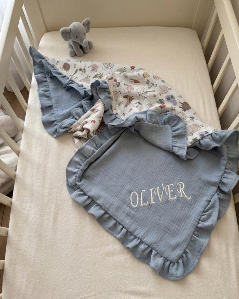 May include: A blue muslin baby blanket with a ruffled edge and the name OLIVER embroidered in white thread. The blanket features a patterned side with illustrations of animals and trees, and a small gray plush elephant toy sits nearby.