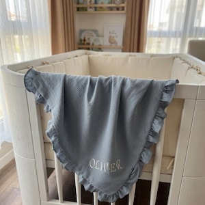 May include: A dusty blue muslin baby blanket with a ruffled edge and the name OLIVER embroidered in white thread. The blanket is draped over the side of a white wooden crib, which features a cream-colored padded bumper lining the interior.
