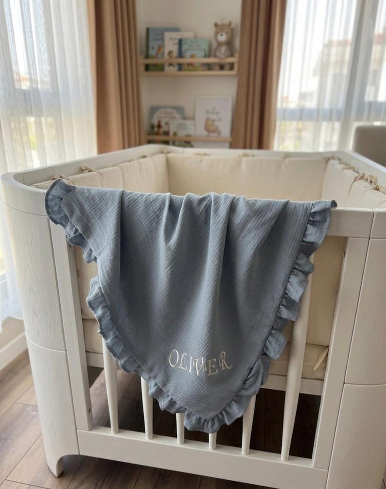 May include: A dusty blue muslin baby blanket with a ruffled edge and the name OLIVER embroidered in white thread. The blanket is draped over the side of a white wooden crib, which features a cream-colored padded bumper lining the interior.