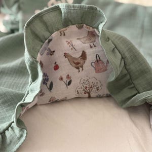 May include: A sage green muslin blanket featuring a ruffled edge and a white underside printed with a pattern of brown chickens, pink watering cans, red apples, and various flowers. The fabric has a soft, crinkled texture throughout the material.