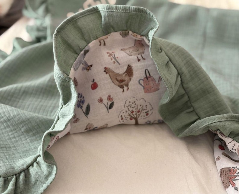 May include: A sage green muslin blanket featuring a ruffled edge and a white underside printed with a pattern of brown chickens, pink watering cans, red apples, and various flowers. The fabric has a soft, crinkled texture throughout the material.
