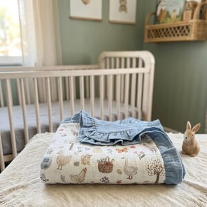 May include: A white muslin baby blanket featuring a repeating pattern of farm animals, including chickens, geese, and a basket of vegetables, finished with a solid blue ruffled border. The fabric has a soft, crinkled texture throughout the design.