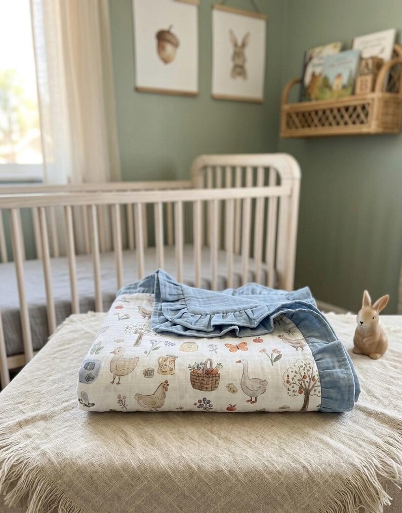 May include: A white muslin baby blanket featuring a repeating pattern of farm animals, including chickens, geese, and a basket of vegetables, finished with a solid blue ruffled border. The fabric has a soft, crinkled texture throughout the design.