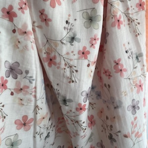 May include: A white fabric featuring a delicate watercolor floral pattern with soft pink, muted purple, and sage green blossoms on thin, dark brown stems. The material has a light, crinkled texture with soft folds and a solid peach-colored border.