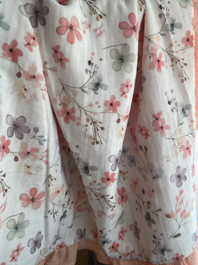 May include: A white fabric featuring a delicate watercolor floral pattern with soft pink, muted purple, and sage green blossoms on thin, dark brown stems. The material has a light, crinkled texture with soft folds and a solid peach-colored border.