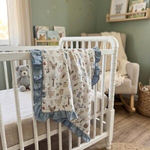 May include: A white muslin baby blanket with a repeating pattern of chickens, trees, carrots, and watering cans, finished with a ruffled blue border. The blanket is draped over the side of a white wooden crib in a nursery setting.