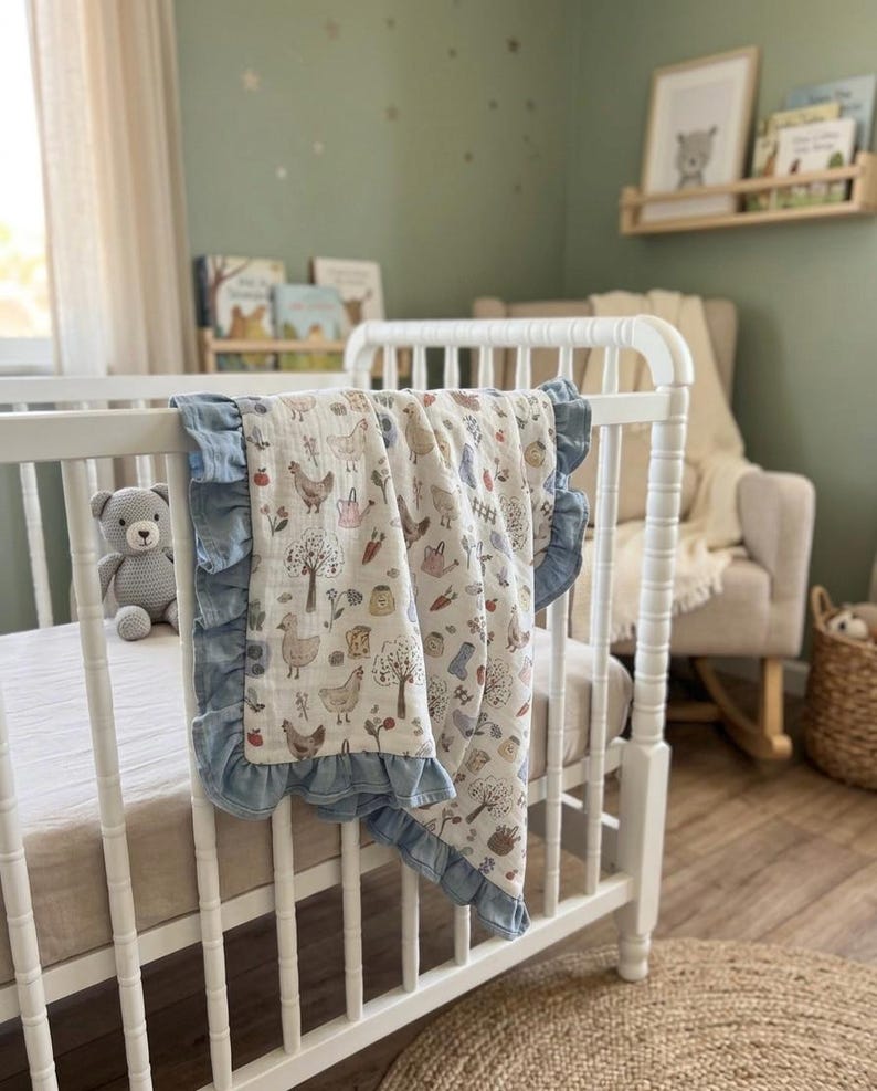 May include: A white muslin baby blanket with a repeating pattern of chickens, trees, carrots, and watering cans, finished with a ruffled blue border. The blanket is draped over the side of a white wooden crib in a nursery setting.