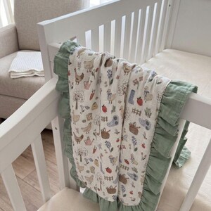 May include: A cream-colored baby blanket featuring a repeating pattern of chickens, ducks, trees, flowers, watering cans, and baskets. The blanket is finished with a ruffled sage green border along the edges and is draped over a white crib rail.